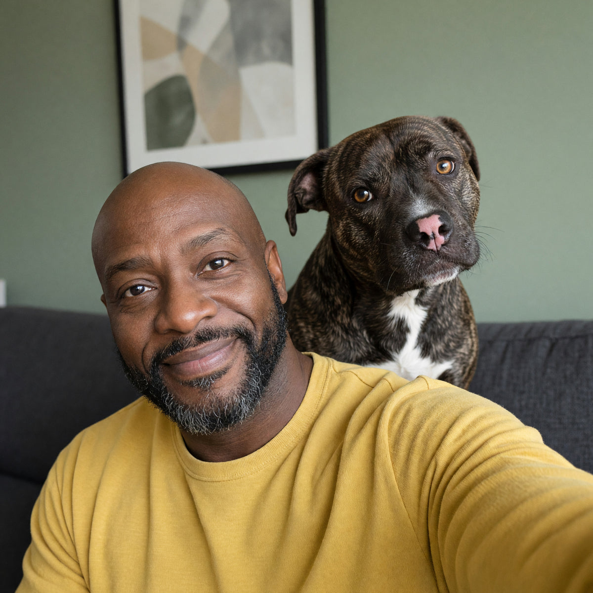 Man in a mustard sweatshirt smiling in a selfie, his brindle pit bull mix sitting on the couch behind him